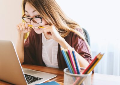 woman biting pencil while sitting on chair in front of computer during daytime